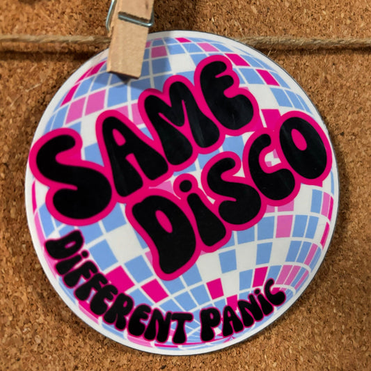 Circular sign with 'Same Disco Different Panic' text on a disco ball background, attached to a cork board.